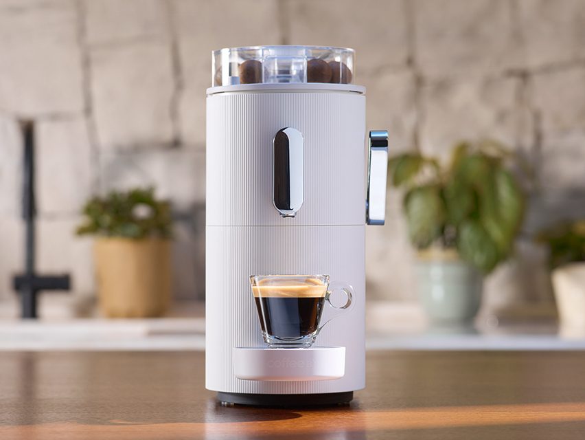 Eine weiße CoffeeB Kaffemaschine steht mit einer gefüllten Glas-Tasse auf einer Küchenzeile.