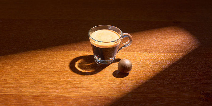 Neben einem angeleuchteten Lungo Glas mit Kaffee liegt ein Coffee Ball.
