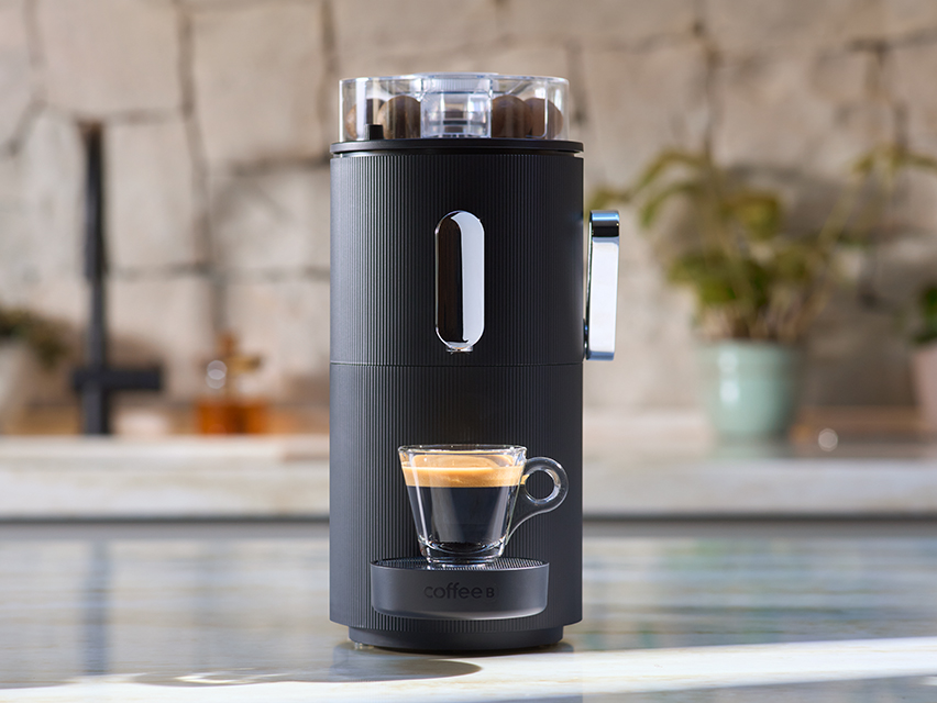 Eine schwarze CoffeeB Kaffeemaschine steht mit einer gefüllten Glas-Tasse auf einer Küchenzeile.
